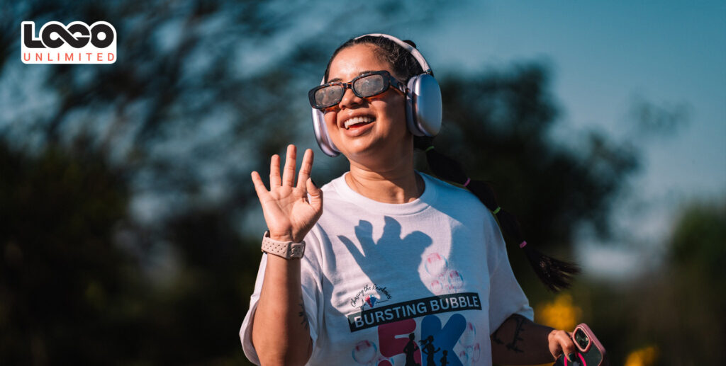 blog-cover-logounlimited-sponsorship-community-seattle-everett-bursting-bubbles-giveaway-event-merch-summer | Logo Unlimited Smiling young woman wearing over-ear headphones and sunglasses waves hello outdoors, LOCO Unlimited logo in the top-left corner.