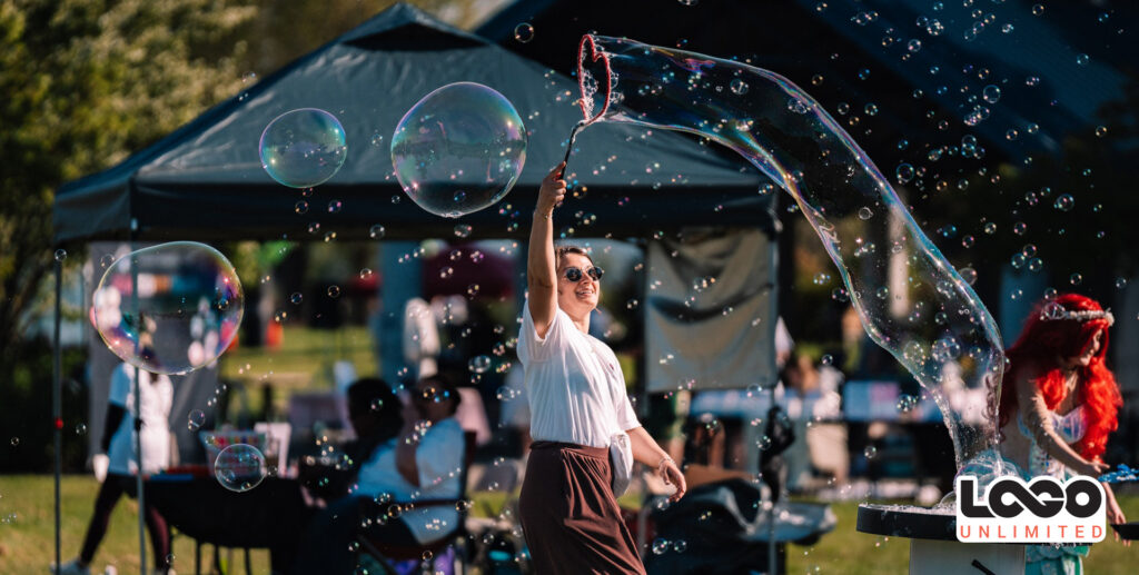blog-cover-logounlimited-sponsorship-community-seattle-everett-bursting-bubbles-giveaway-event-merch-summer-medal-running-5k-eventseason-donation | Logo Unlimited Woman with a bubble wand creating large bubbles at an outdoor festival, smiling in sunglasses under a canopy.