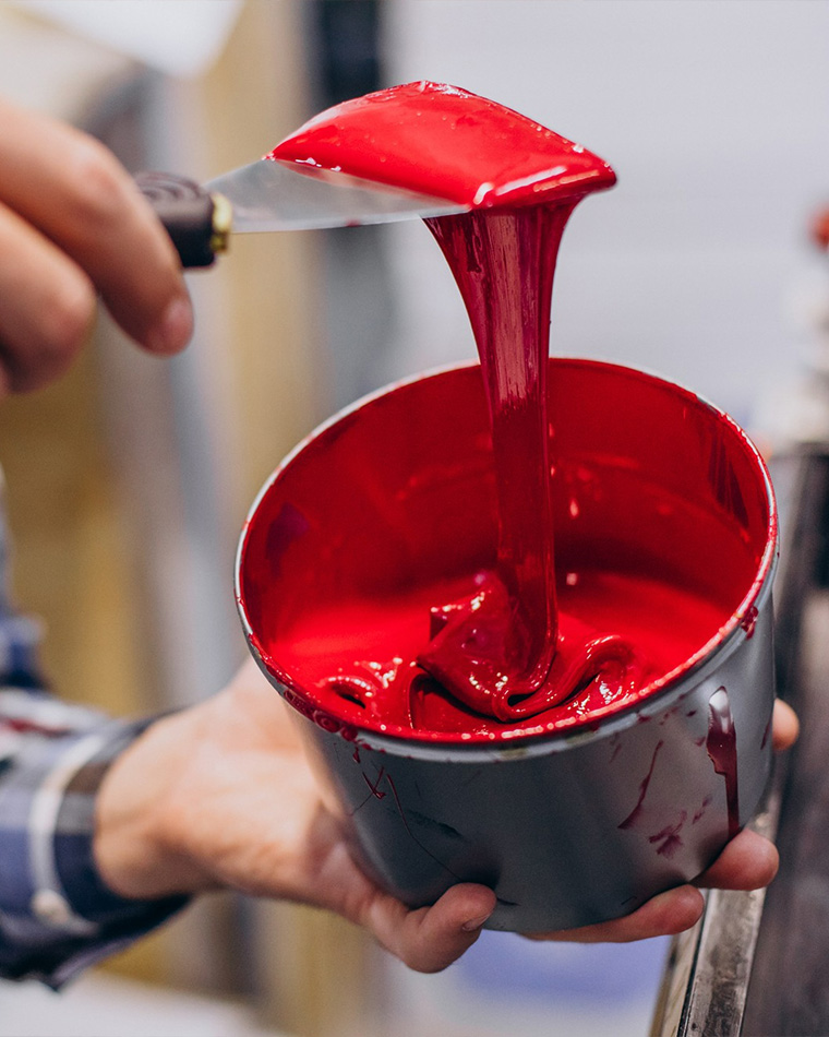 Close-up of a hand holding a can while a bright red paint is being spread with a putty knife, creating a glossy stream of paint into the can's rim.