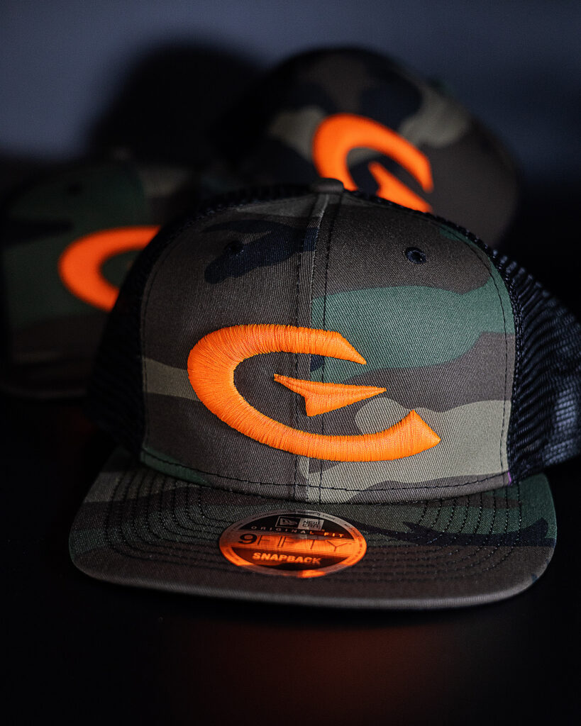 Close-up of a camouflage baseball cap with bright orange embroidered letter logo and matching orange eyelets, shallow depth of field.