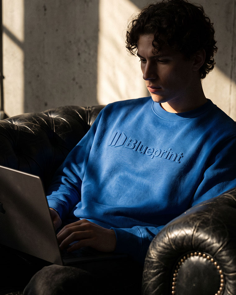 Young man in a blue Blueprint sweatshirt typing on a laptop while seated on a leather couch. (informative)