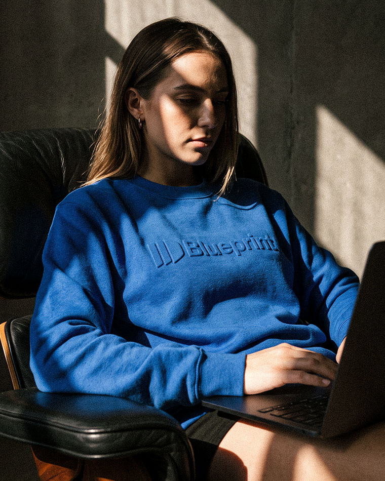 Woman seated in a dark chair, wearing a blue sweatshirt with an embossed logo, typing on a laptop in sunlight.