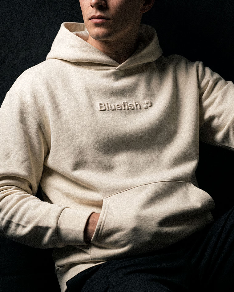 Person in a cream hoodie with an embroidered logo on the chest, seated against a dark background.