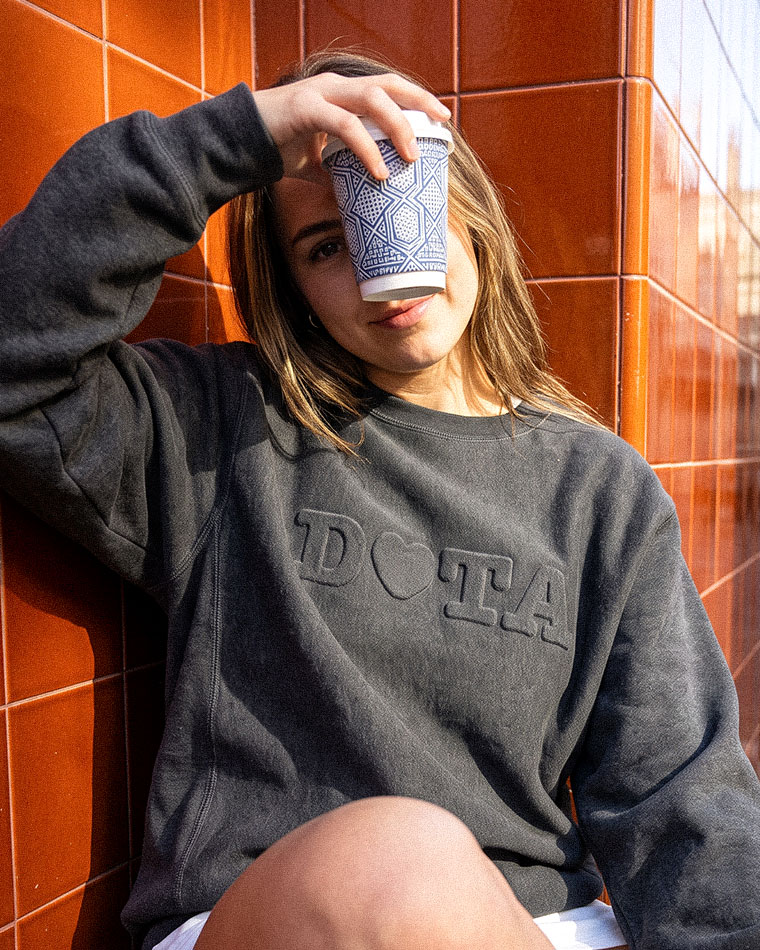 Young woman leaning against orange tiled wall, holding a blue patterned cup over her forehead in a casual pose