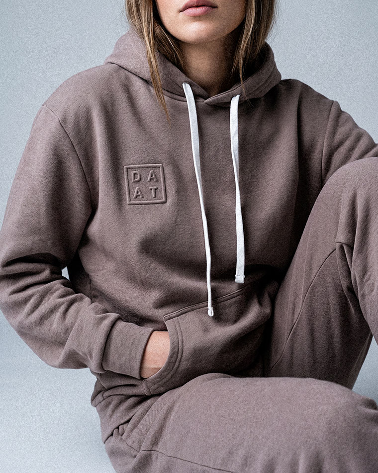 Person wearing a taupe hoodie with white drawstrings; logo patch on chest reads 'DAAT.'
