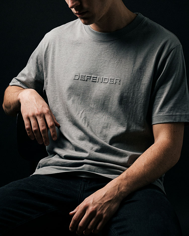 Person wearing a light gray T-shirt with an embossed 'DEFENDER' logo, seated against a dark background.