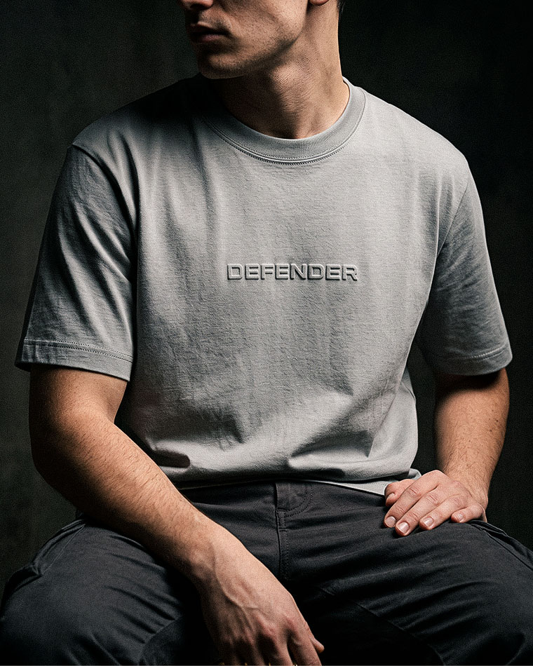Man wearing a light gray T-shirt with an embossed 'DEFENDER' logo, seated against a dark background.