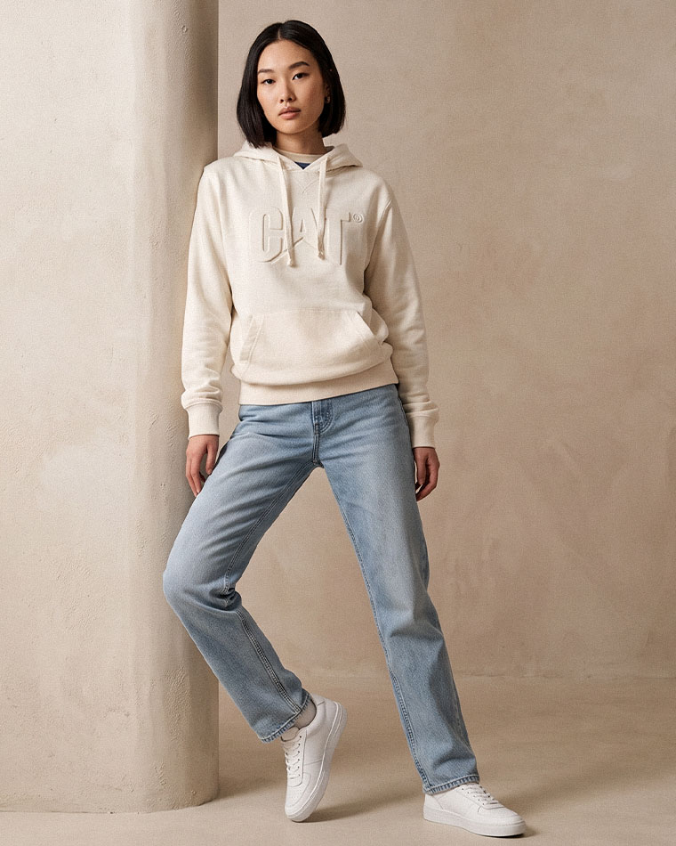 Woman in a cream hoodie with a raised logo, light-wash jeans, and white sneakers leaning against a column in a neutral studio setting.