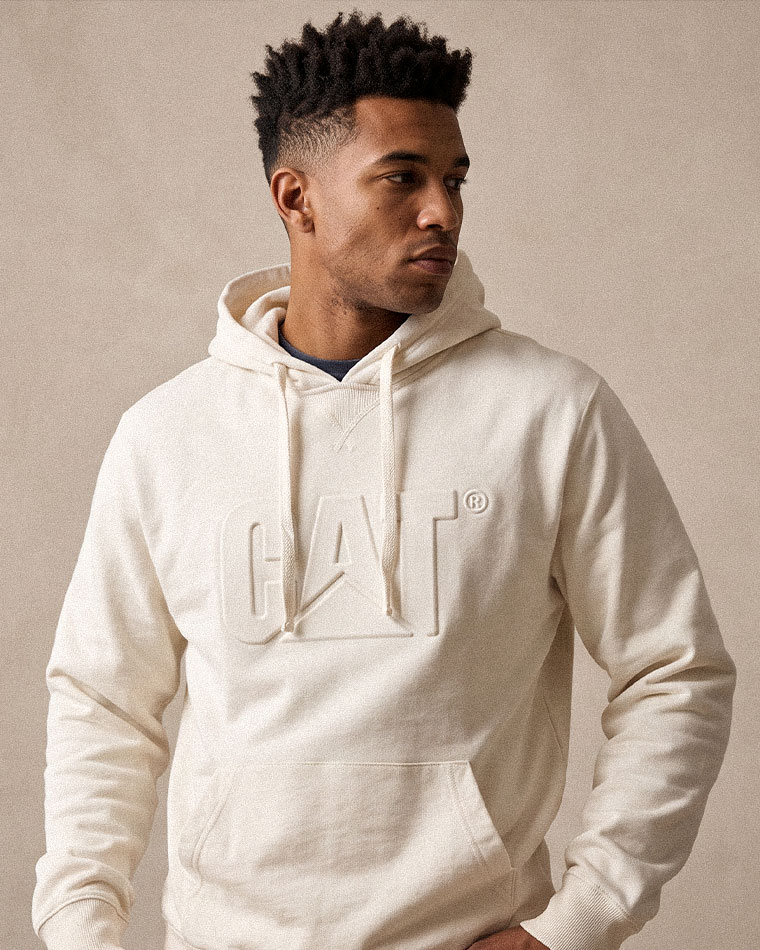 Man modeling a cream hoodie with a large embossed CAT logo on the chest, looking to the right against a neutral background.