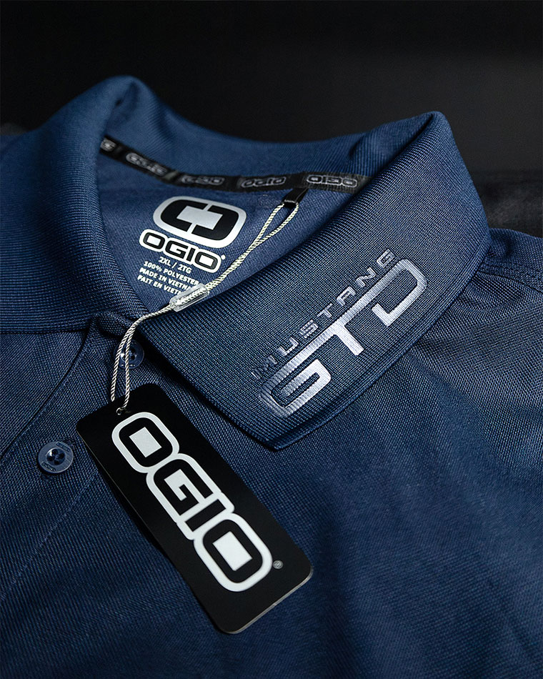 Blue polo shirt close-up showing OGIO tag and the OGIO logo on the collar patch, with a hanging brand tag.