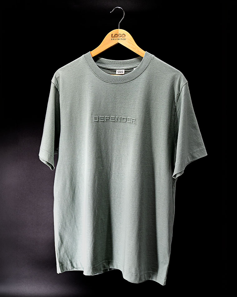 Grey short-sleeve T-shirt on a wooden hanger against a dark background; has 'DEFENDER' embossed on the chest.