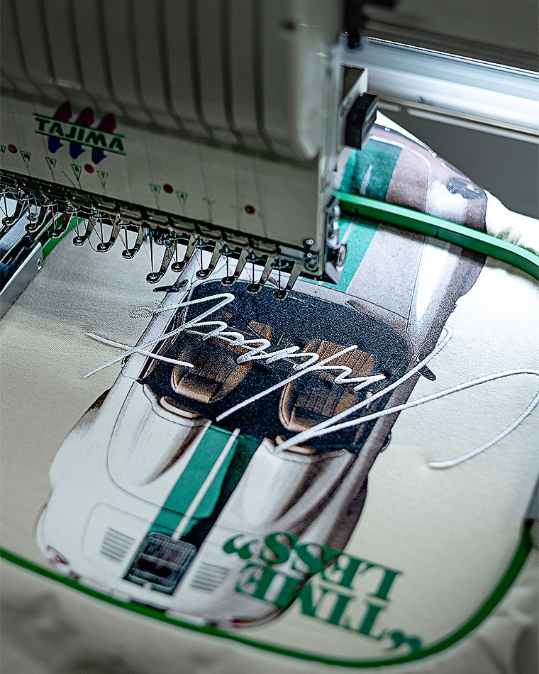 Close-up of an embroidery machine stitching a white cursive design onto fabric, with Tajima branding visible.